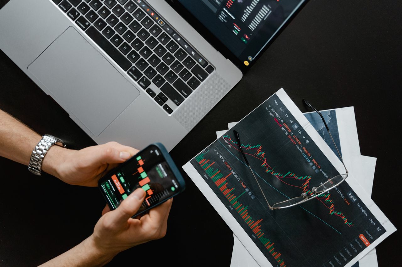 services-03 Hands using smartphone beside laptop with stock charts, showcasing digital trading.